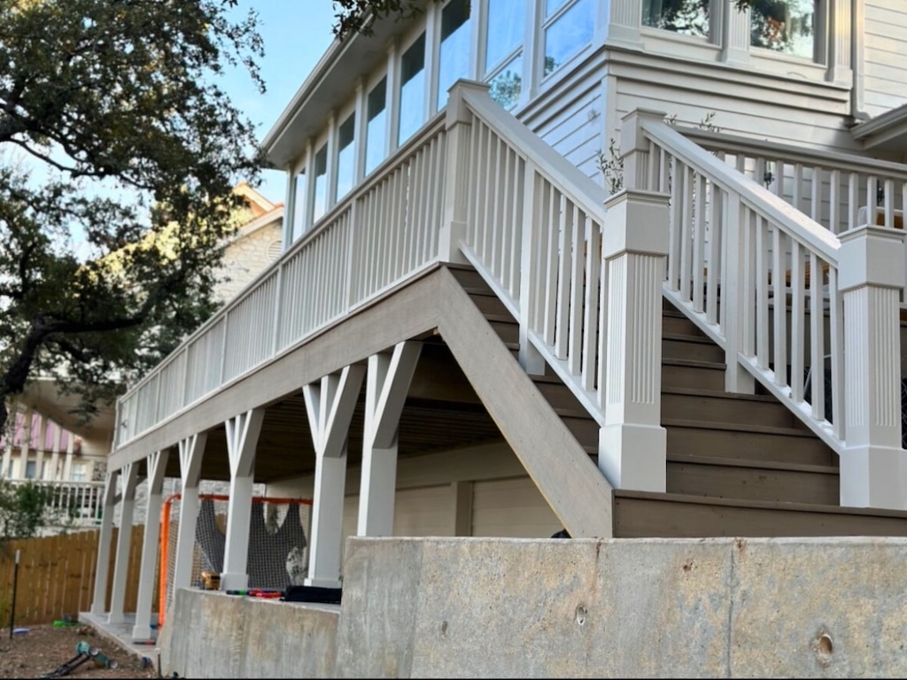 Elevated white deck with wooden stairs and railing, attached to a house. Overlooking tall trees and a sunny yard, creating a serene atmosphere.