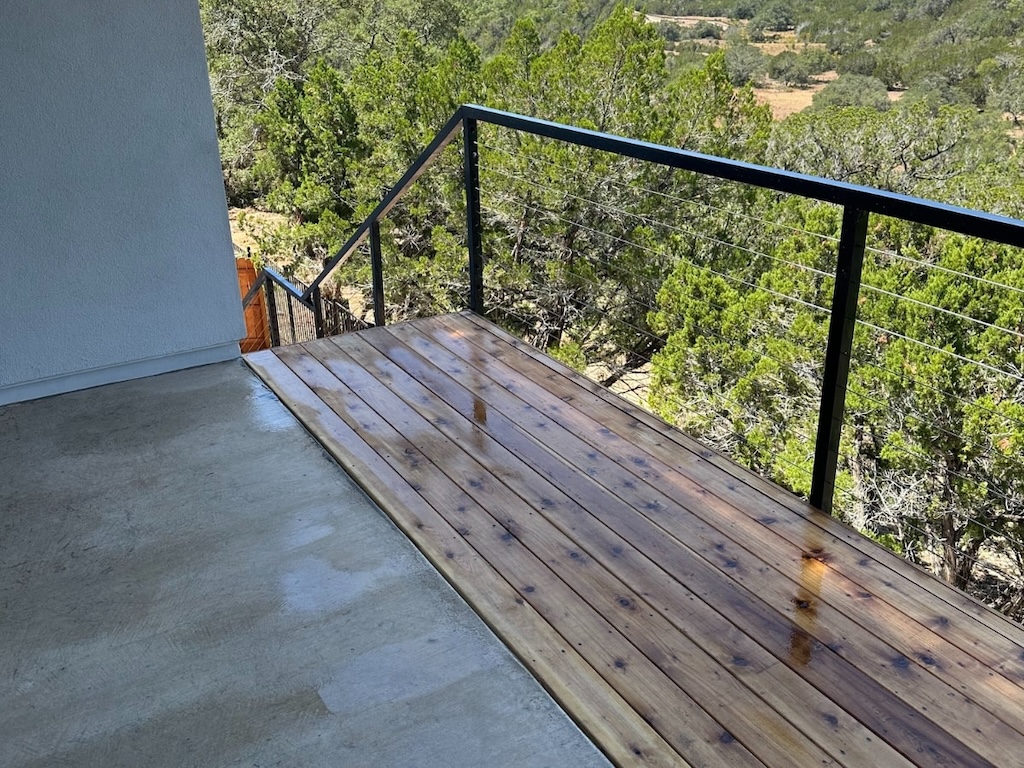 A wet wooden deck leads to a railing overlooking a lush green forest. The scene conveys a serene, natural atmosphere under a sunny sky.