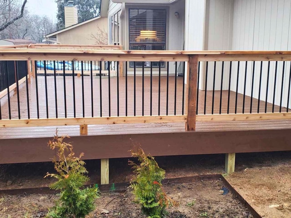 A newly built wooden deck with light brown planks and black metal railings surrounded by a small garden with young shrubs. A cozy house is visible behind.
