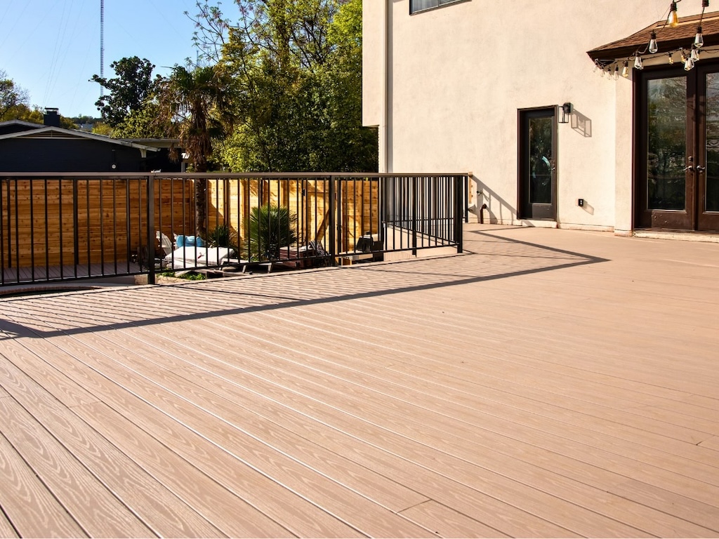 spacious wooden deck with black railing is attached to a cream-colored house. Trees and a wooden fence surround a cozy patio area. Bright, sunny day.