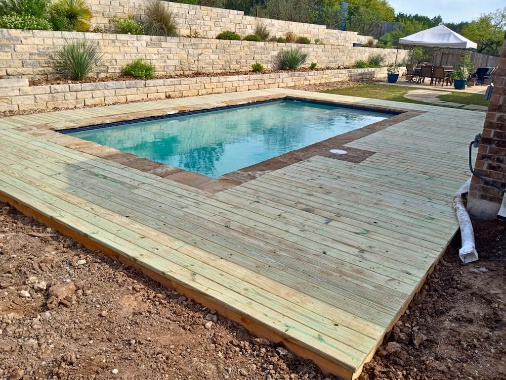 Backyard scene showing a rectangular pool with clear water, surrounded by a wooden deck. Terraced stone walls with plants add a natural vibe. A canopy and table set are in the background.