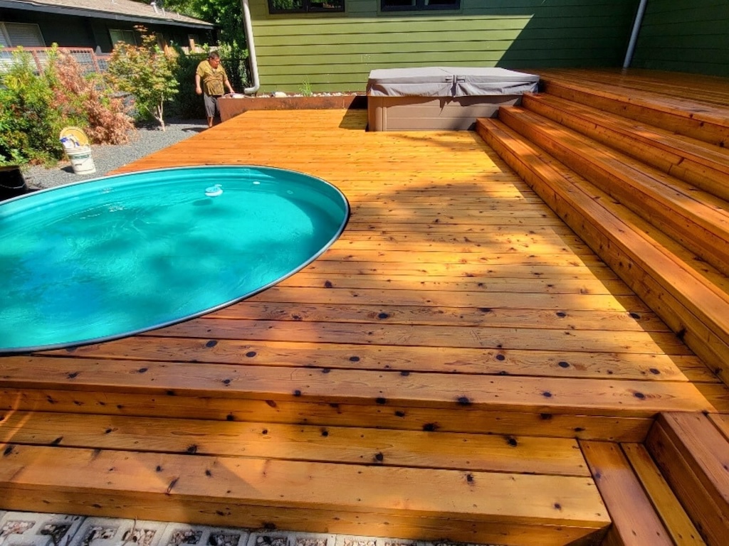 A sunlit wooden deck with an oval pool and multi-level steps. A man tends plants in the corner, creating a serene backyard atmosphere.