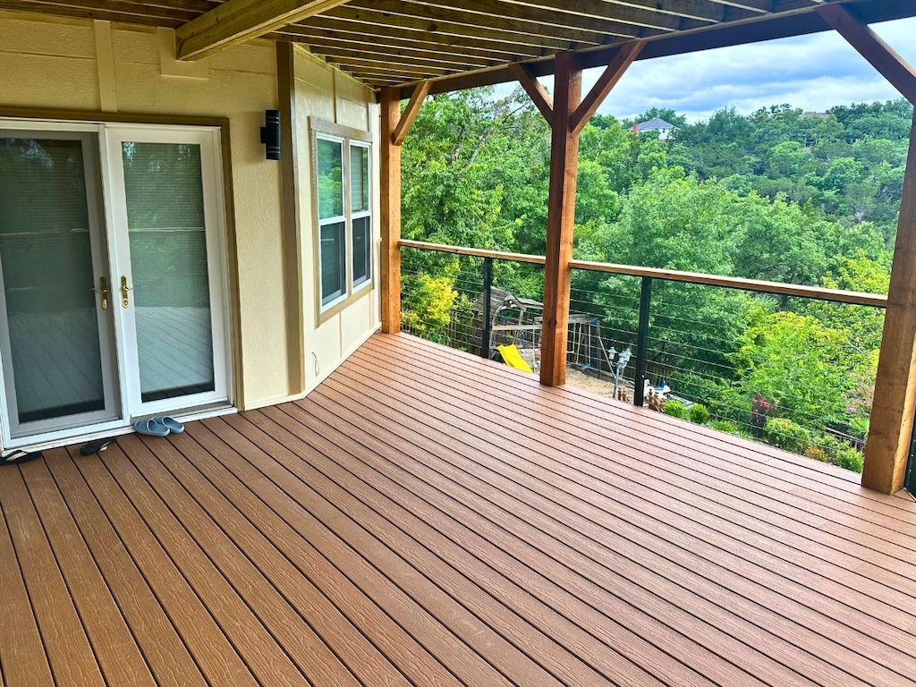 Covered wooden deck with railing, overlooking lush green trees and a distant house. Glass doors and slippers suggest relaxation. Bright, serene atmosphere.