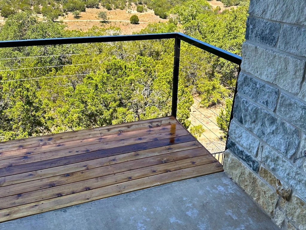Small wooden balcony with metal railing, overlooking a lush forest under a clear blue sky. A stone wall borders the right side, creating a serene, rustic atmosphere.