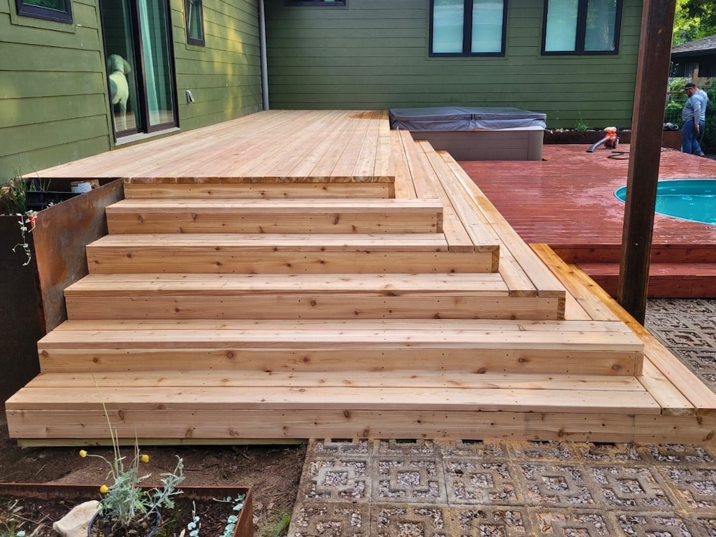 Wide wooden deck with multiple steps leads up to a green house. A smaller deck surrounds a hot tub and pool. Calm atmosphere with greenery.