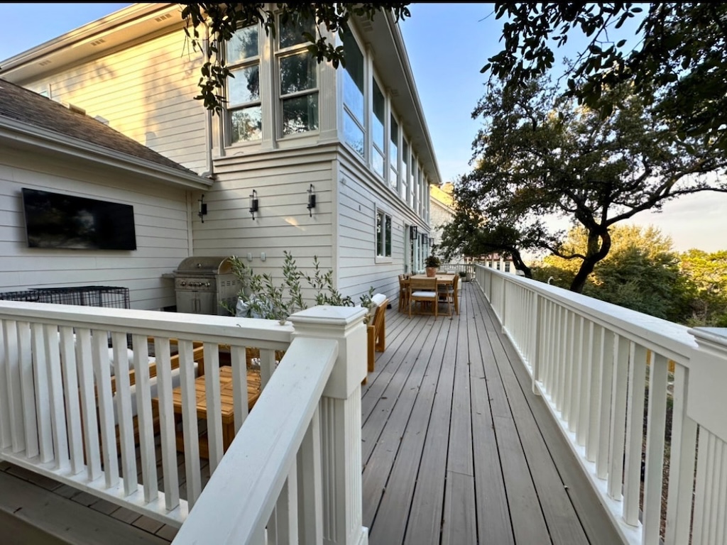 Wooden deck with white railing attached to a house, featuring outdoor furniture, a mounted TV, and a grill. Trees create a serene, inviting atmosphere.