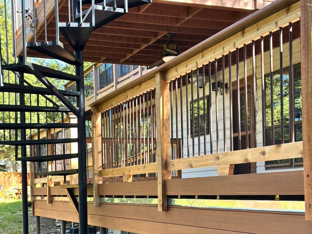 Outdoor spiral staircase beside a wooden deck railing. Sunlight casts shadows, creating a tranquil, natural atmosphere with trees in the background.