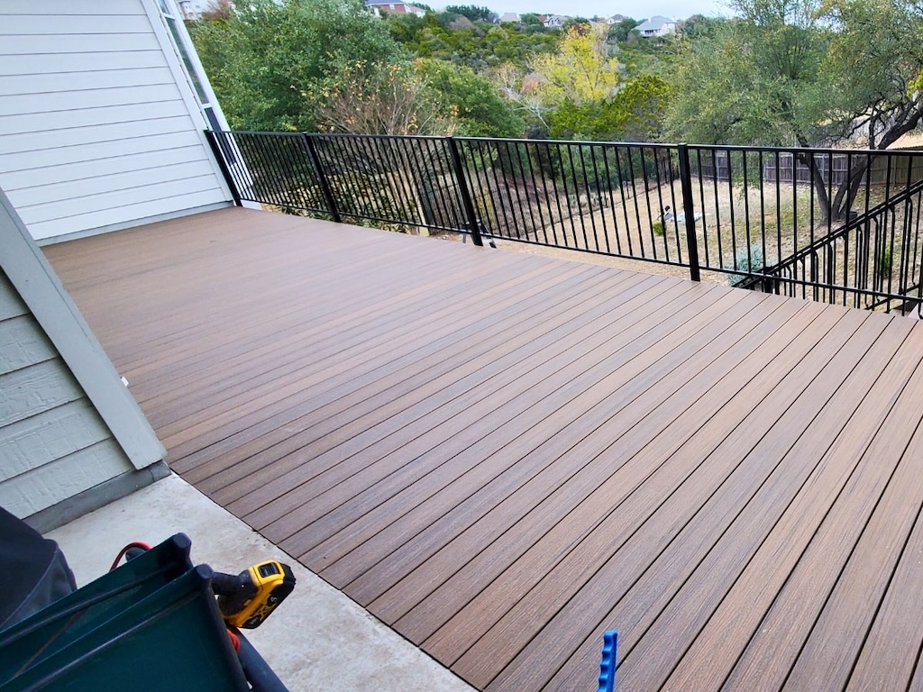 A newly built wooden deck with brown planks, bordered by a black metal railing, overlooks a green, tree-filled landscape, conveying a serene outdoor setting.