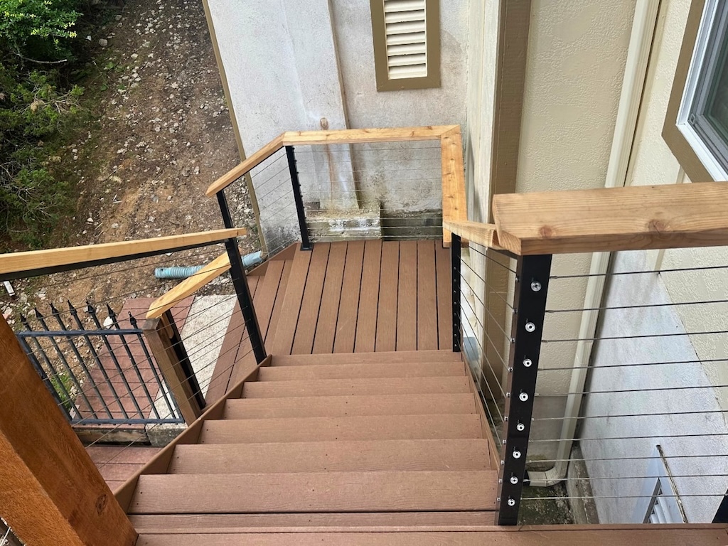 Wooden steps with sleek metal cable railings descend alongside a beige wall. The outdoor setting includes plants and a stone path below.