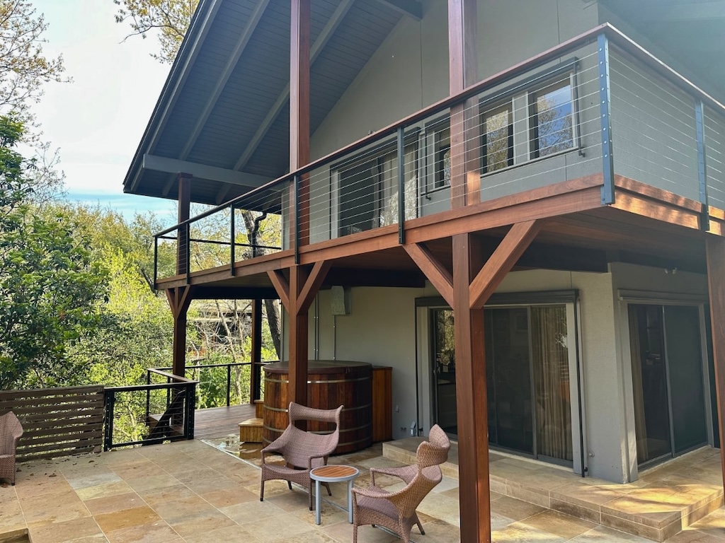 Two-story house with wooden beams and metal railing, featuring a large balcony. Stone patio below has wicker chairs and a hot tub, surrounded by green trees.