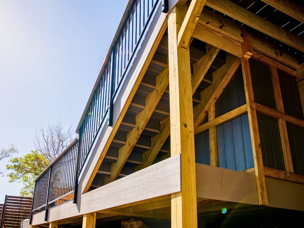 Wooden deck with black railings shown from below, highlighting sturdy beams and open stairs. Bright sky and trees suggest a sunny day outdoors.