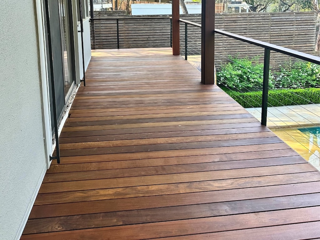 Newly built wooden deck with a smooth finish, adjacent to a modern house. The deck overlooks a lush garden with green hedges and a pool, evoking tranquility.