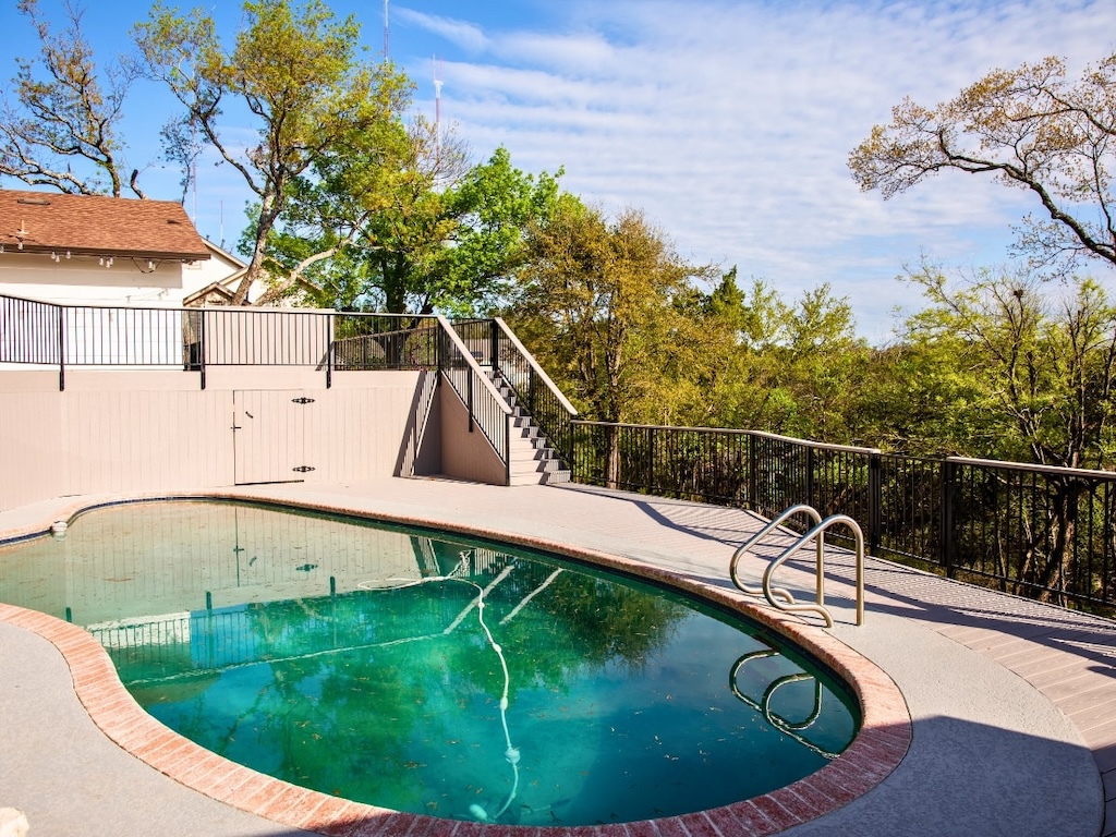 Outdoor pool area with oval-shaped pool surrounded by a concrete deck, brick trim, and metal railings. Trees and a house in the background under a sunny sky.