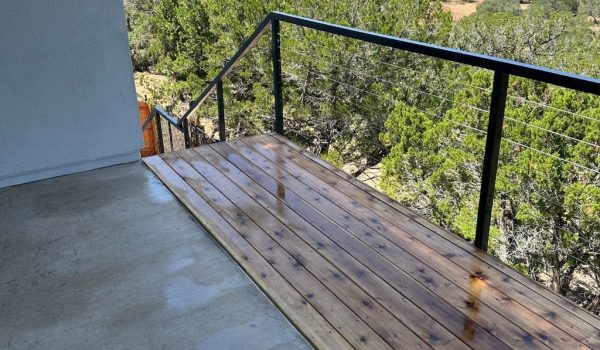 A wet wooden deck leads to a railing overlooking a lush green forest. The scene conveys a serene, natural atmosphere under a sunny sky.