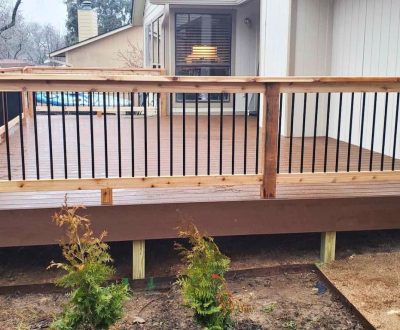 A newly built wooden deck with light brown planks and black metal railings surrounded by a small garden with young shrubs. A cozy house is visible behind.