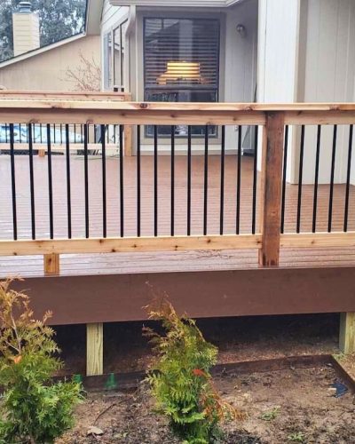A newly built wooden deck with light brown planks and black metal railings surrounded by a small garden with young shrubs. A cozy house is visible behind.