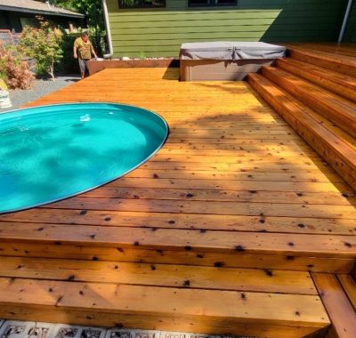 A sunlit wooden deck with an oval pool and multi-level steps. A man tends plants in the corner, creating a serene backyard atmosphere.