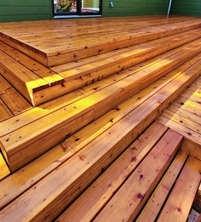 stained wooden outdoor deck with wide, tiered steps, basking in warm sunlight, creating a welcoming, natural ambiance against a green wall backdrop.