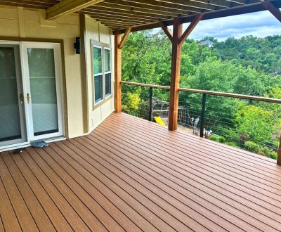 Covered wooden deck with railing, overlooking lush green trees and a distant house. Glass doors and slippers suggest relaxation. Bright, serene atmosphere.