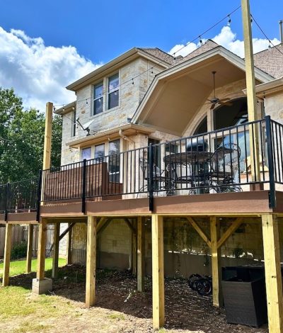 Stone house with a raised wooden deck supported by beams, featuring black metal railings and patio furniture. Sunny day with blue sky and clouds.