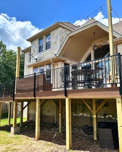 Stone house with a raised wooden deck supported by beams, featuring black metal railings and patio furniture. Sunny day with blue sky and clouds.