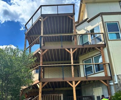 Outdoor staircase with black metal railings leading to a rooftop deck on a stone house. The setting is sunny with trees and an umbrella in the background.