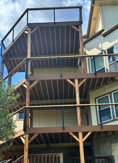 Outdoor staircase with black metal railings leading to a rooftop deck on a stone house. The setting is sunny with trees and an umbrella in the background.