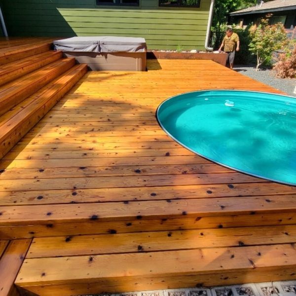 Wooden deck with an above-ground pool embedded, surrounded by a sunlit, wooden platform. A person stands by a green building, creating a relaxed outdoor vibe.