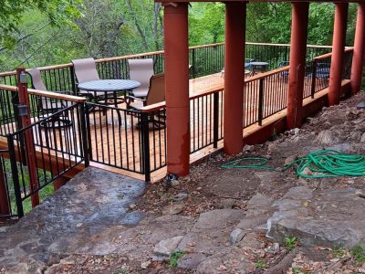 Wooden deck with metal railings, table, and chairs overlooking lush green trees. Earthy tones and hoses on rocky ground suggest a serene, rustic setting.