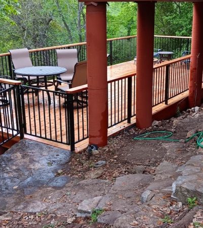 Wooden deck with metal railings, table, and chairs overlooking lush green trees. Earthy tones and hoses on rocky ground suggest a serene, rustic setting.