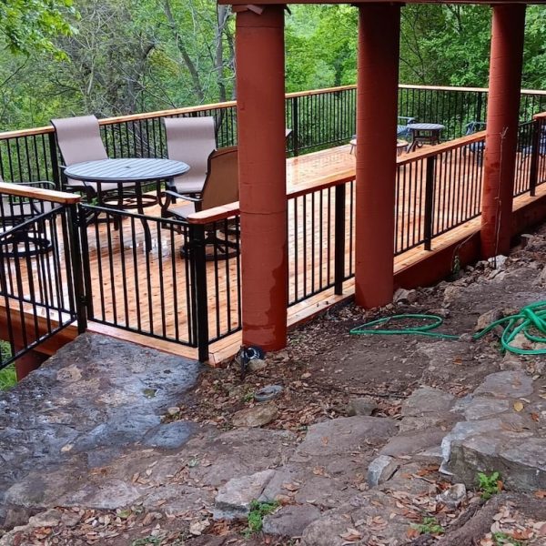 Wooden deck with metal railings, table, and chairs overlooking lush green trees. Earthy tones and hoses on rocky ground suggest a serene, rustic setting.