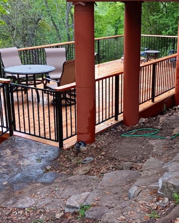 Wooden deck with metal railings, table, and chairs overlooking lush green trees. Earthy tones and hoses on rocky ground suggest a serene, rustic setting.
