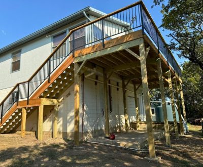Elevated wooden deck with black railings attached to a light gray house. Sunlight casts shadows on the structure, creating a warm, serene atmosphere.