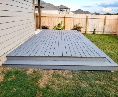 A simple gray composite deck extends from a house onto a grassy backyard, bordered by a beige wooden fence. The sky above is partly cloudy, suggesting a calm, neutral atmosphere.