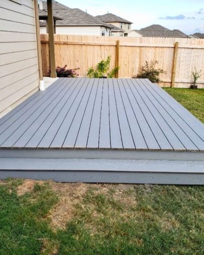 A simple gray composite deck extends from a house onto a grassy backyard, bordered by a beige wooden fence. The sky above is partly cloudy, suggesting a calm, neutral atmosphere.