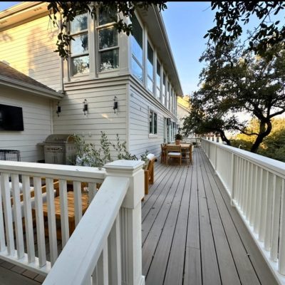 Wooden deck with white railing attached to a house, featuring outdoor furniture, a mounted TV, and a grill. Trees create a serene, inviting atmosphere.