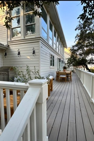 Wooden deck with white railing attached to a house, featuring outdoor furniture, a mounted TV, and a grill. Trees create a serene, inviting atmosphere.