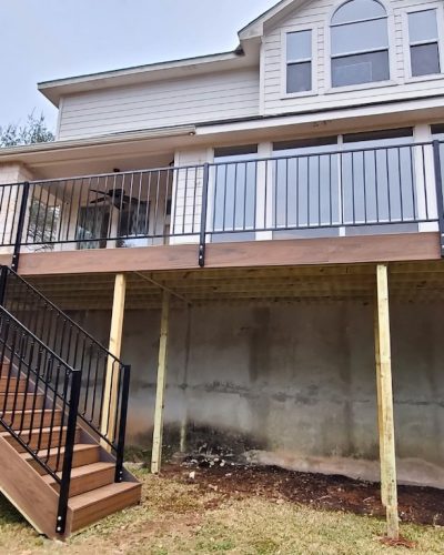 Two-story house with large upper deck featuring black railings and wooden stairs. Overcast sky, trees in background, conveying a calm suburban setting.
