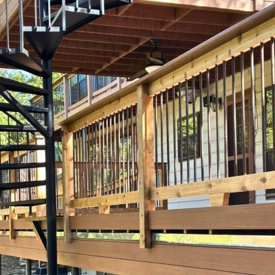 Outdoor spiral staircase beside a wooden deck railing. Sunlight casts shadows, creating a tranquil, natural atmosphere with trees in the background.