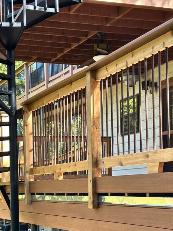 Outdoor spiral staircase beside a wooden deck railing. Sunlight casts shadows, creating a tranquil, natural atmosphere with trees in the background.