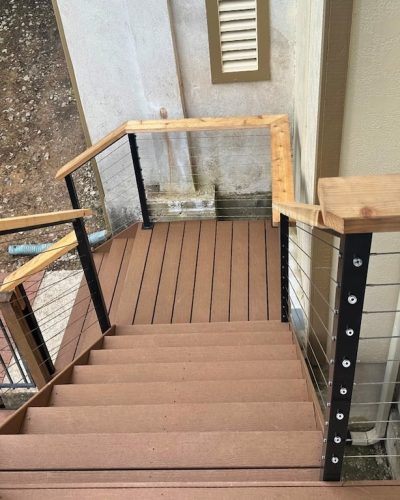 Wooden steps with sleek metal cable railings descend alongside a beige wall. The outdoor setting includes plants and a stone path below.