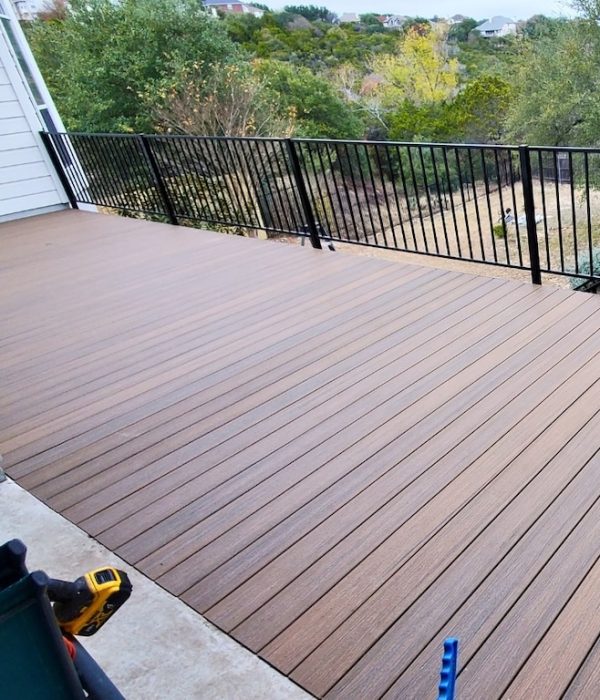 A newly built wooden deck with brown planks, bordered by a black metal railing, overlooks a green, tree-filled landscape, conveying a serene outdoor setting.