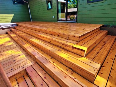 Outdoor wooden deck with wide, polished steps leading to a green house wall. Sunlight highlights the warm, rich tones of the wooden planks.