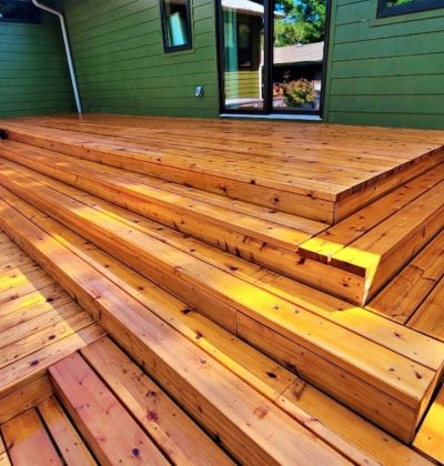 Outdoor wooden deck with wide, polished steps leading to a green house wall. Sunlight highlights the warm, rich tones of the wooden planks.