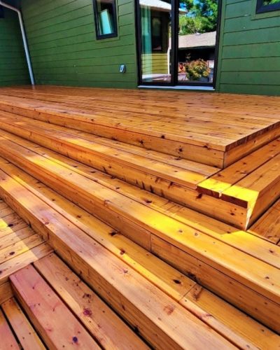 Outdoor wooden deck with wide, polished steps leading to a green house wall. Sunlight highlights the warm, rich tones of the wooden planks.