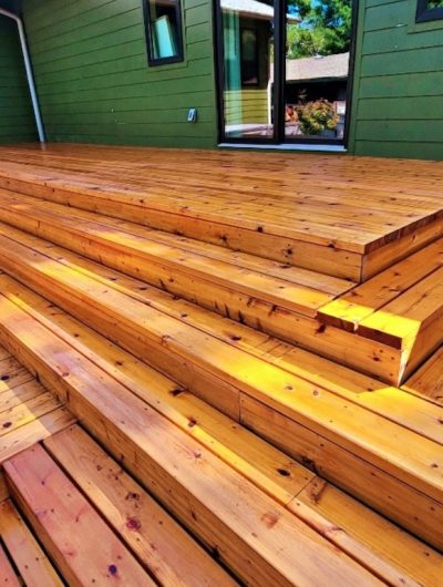 Outdoor wooden deck with wide, polished steps leading to a green house wall. Sunlight highlights the warm, rich tones of the wooden planks.