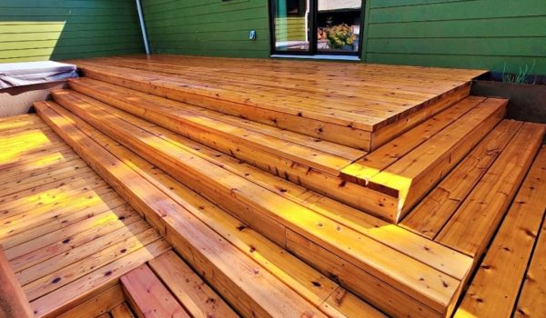 Outdoor wooden deck with wide, polished steps leading to a green house wall. Sunlight highlights the warm, rich tones of the wooden planks.