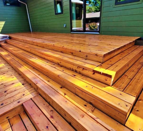 Outdoor wooden deck with wide, polished steps leading to a green house wall. Sunlight highlights the warm, rich tones of the wooden planks.