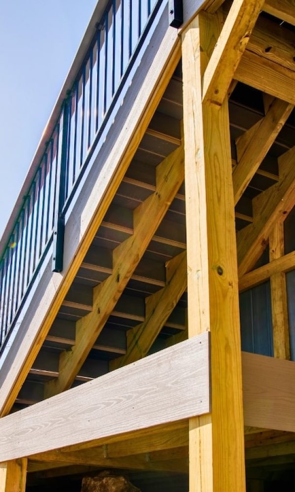 Wooden deck with black railings shown from below, highlighting sturdy beams and open stairs. Bright sky and trees suggest a sunny day outdoors.