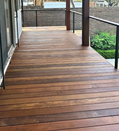 Newly built wooden deck with a smooth finish, adjacent to a modern house. The deck overlooks a lush garden with green hedges and a pool, evoking tranquility.